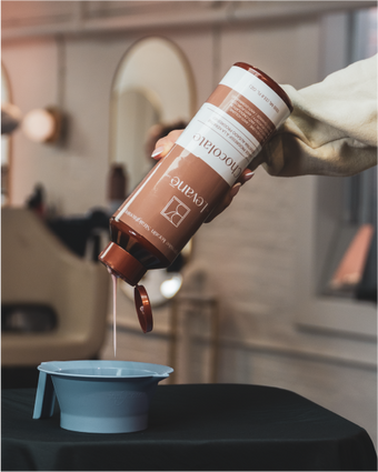 A hand pours the contents of a bottle of chocolate keratin into a container.